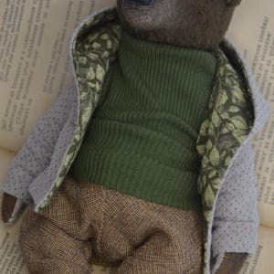 A brown teddy bear wearing a green sweater and brown pants is lying on an open book.