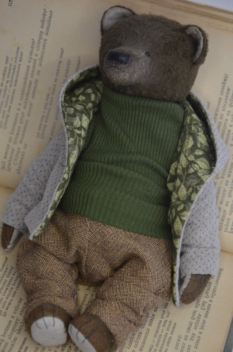 A brown teddy bear wearing a green sweater and brown pants is lying on an open book.