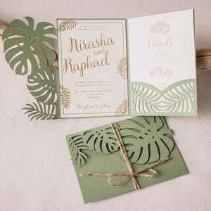 Peut inclure: Un ensemble de faire-part de mariage vert sauge avec des motifs de feuilles de monstera découpés au laser. La carte ouverte affiche les noms Nirasha et Raphael en lettres dorées, et l'enveloppe pliée est nouée avec une ficelle brune.