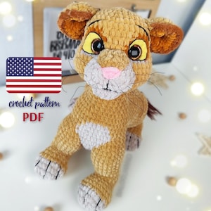 May include: A plush lion toy made of golden-brown yarn with a light gray muzzle, chest, and paw pads. The lion has large, dark, glossy eyes with yellow irises, a pink nose, and black whiskers. The text "crochet pattern PDF" is visible on the left.