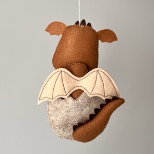 May include: A brown felt dragon hanging ornament with beige wings and a brown tail. The dragon has a stitched design on its wings and tail.