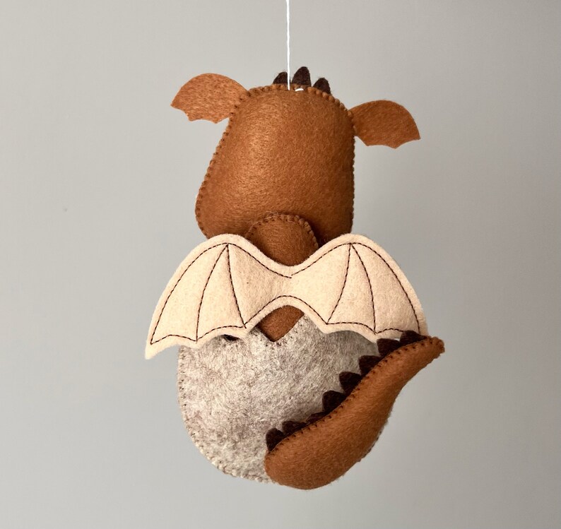 May include: A brown felt dragon hanging ornament with beige wings and a brown tail. The dragon has a stitched design on its wings and tail.