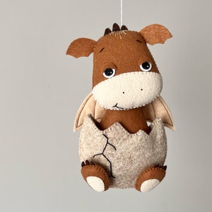 May include: A brown felt dragon with a beige felt egg, hatching from the egg. The dragon has black eyes and a stitched smile.