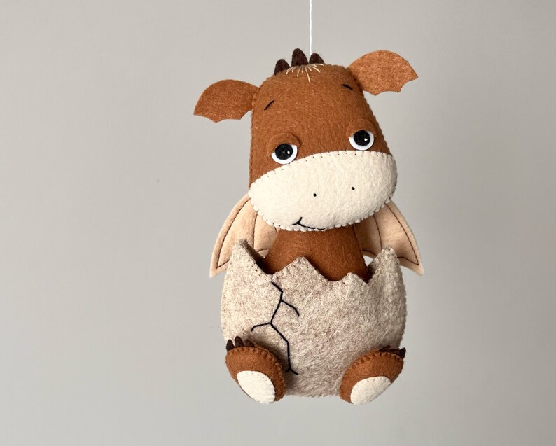 May include: A brown felt dragon with a beige felt egg, hatching from the egg. The dragon has black eyes and a stitched smile.