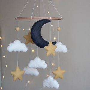 Può includere: Un mobile per bambini in legno con una luna crescente in feltro nero, nuvole in feltro bianco e stelle in feltro dorate. Il mobile è appeso a un anello di legno.