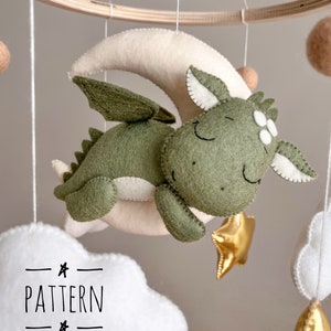 Può includere: Un drago verde in feltro con occhi bianchi e pancia bianca sta dormendo su una luna crescente bianca in feltro. Il drago tiene una stella dorata in feltro. Il drago fa parte di un mobile con nuvole bianche in feltro e una stella dorata in feltro.