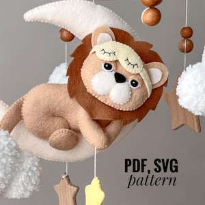 May include: A felt lion with a sleep mask on its eyes is lying on a white crescent moon. The moon is attached to a wooden mobile with white fluffy clouds, wooden stars, and brown wooden beads. The text "PDF, SVG pattern" is printed below the mobile.