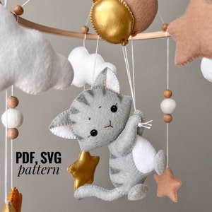 Può includere: Un mobile in legno con un gatto grigio e bianco che tiene una stella dorata, nuvole, stelle e palloncini. Il mobile è fatto di feltro ed è un modello fai-da-te per un mobile per bambini.