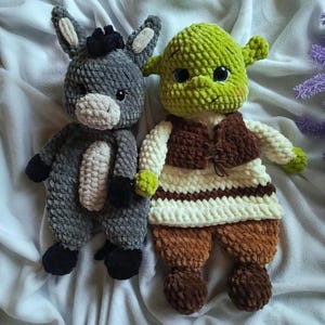 Op de afbeelding: Twee handgemaakte gehaakte poppen: een grijze ezel met zwarte hoeven en een Shrek-pop. De Shrek-pop is groen met een bruin vest, een wit-bruin gestreept shirt en een bruine broek. Beide poppen staan op een wit oppervlak.