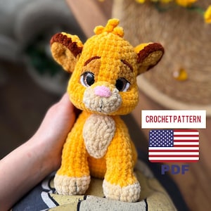 May include: A crocheted lion cub toy in shades of yellow, tan, and brown. The lion cub has large, expressive eyes, a pink nose, and a fluffy mane. The image includes text that says "CROCHET PATTERN" and a US flag.