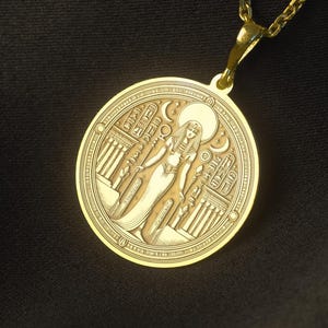 May include: A gold-colored pendant necklace featuring a circular charm with an engraved design of a figure and architectural elements. The pendant hangs from a delicate chain. The background is a dark, textured fabric.