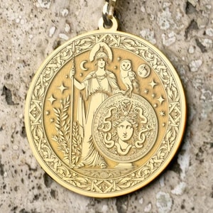 May include: Gold-colored pendant necklace featuring a detailed circular medallion. The medallion depicts a classical figure with an owl, shield, and stars. The pendant hangs from a delicate gold chain, creating an elegant accessory.
