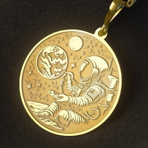 May include: Gold necklace with a round pendant featuring an astronaut blowing bubbles in space. The pendant has a detailed design of the astronaut, a planet, and stars.