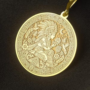May include: A gold-toned circular pendant necklace featuring an intricate engraved design of a figure wearing a feathered headdress, surrounded by floral motifs, birds, and a crescent moon, all enclosed within a geometric patterned border.