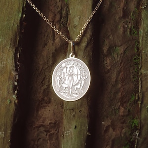 May include: A silver pendant necklace with a circular charm depicting a woman standing in a forest, with a crescent moon and stars above her. The charm is surrounded by a border of leaves and flowers.