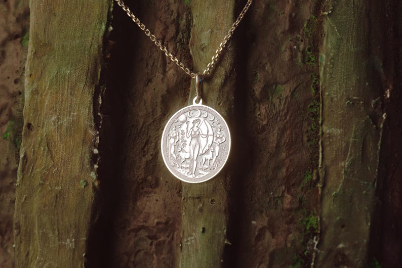 May include: A silver pendant necklace with a circular charm depicting a woman standing in a forest, with a crescent moon and stars above her. The charm is surrounded by a border of leaves and flowers.