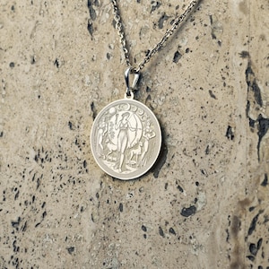 May include: Silver necklace with a round pendant featuring a detailed design of a woman with a bow and arrow, surrounded by a crescent moon and stars.