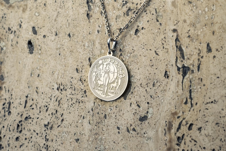 May include: Silver necklace with a round pendant featuring a detailed design of a woman with a bow and arrow, surrounded by a crescent moon and stars.