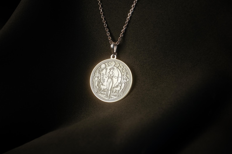 May include: A silver necklace with a round pendant featuring a detailed engraving of a woman standing in a forest with a crescent moon above her head.