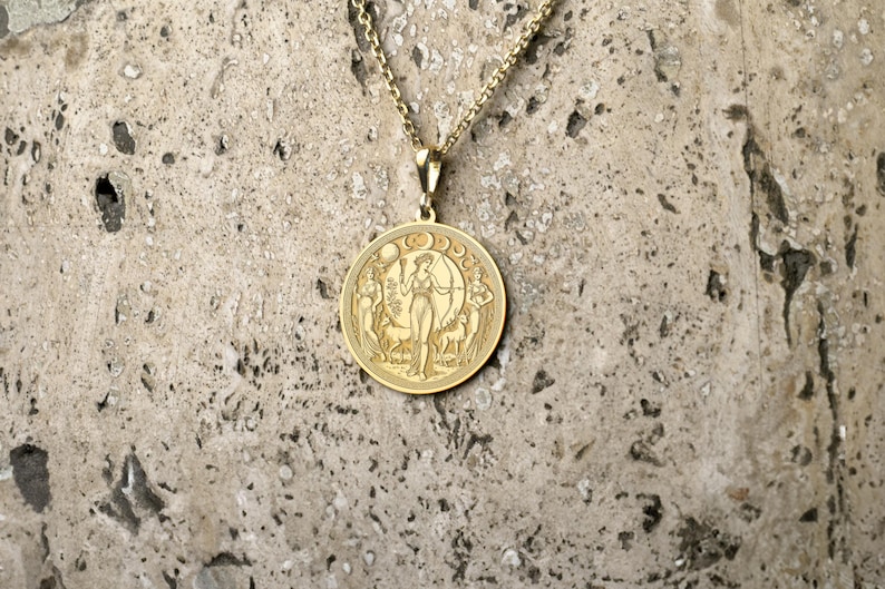 May include: A gold necklace with a round pendant featuring a detailed engraving of a woman standing in a forest with a crescent moon above her head.