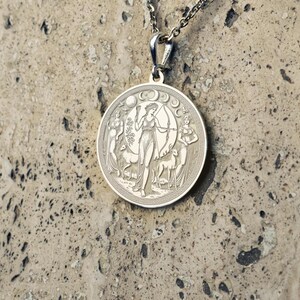 May include: Silver pendant necklace featuring a circular design with an engraved image of a woman holding a bow and arrow, surrounded by two deer and crescent moons.