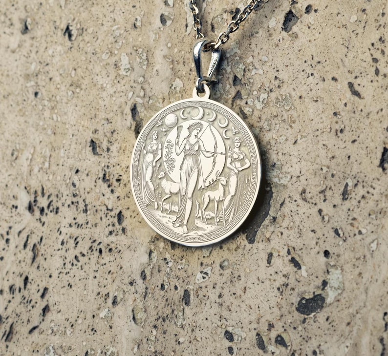 May include: Silver pendant necklace featuring a circular design with an engraved image of a woman holding a bow and arrow, surrounded by two deer and crescent moons.