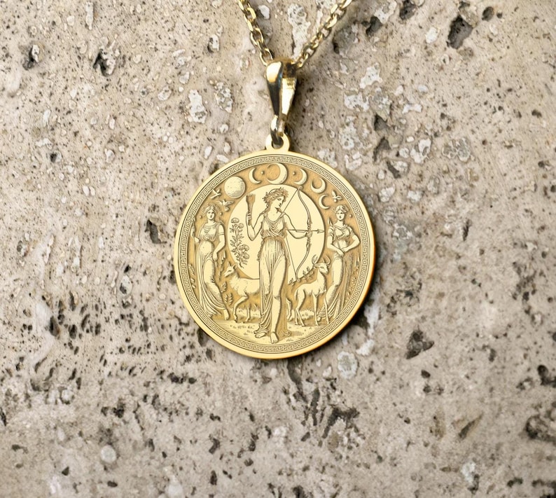 May include: A gold pendant necklace with a round, engraved medallion featuring a woman with a bow and arrow, surrounded by crescent moons and other figures.