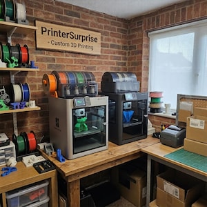 May include: A workshop with two 3D printers and multiple spools of filament in various colours. The printers sit on a wooden table, with a sign reading "PrinterSurprise - Custom 3D Printing" on the brick wall. Boxes and a label printer are also visible.