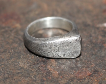May include: A silver ring with a wide, flat band. The band has a textured, hammered finish.