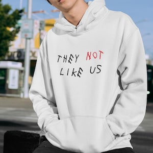 May include: White hooded sweatshirt with the phrase "THEY NOT LIKE US" written in black and red on the front.