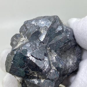 Può includere: Un campione di minerale grigio scuro con una struttura cristallina. La superficie del minerale ha una lucentezza metallica, che riflette la luce. Il minerale è tenuto da una mano guantata. L'aspetto generale suggerisce una gemma o un minerale naturale grezzo.