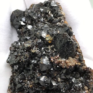 Puede incluir: Un cúmulo de cristales negros, brillantes y afilados sobre una matriz marrón. Los cristales están densamente agrupados y tienen un brillo metálico.