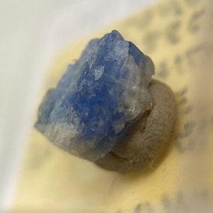 May include: A small, blue, crystalline mineral specimen on a grey background.