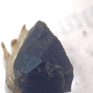 Puede incluir: Un cristal de color gris oscuro, de forma irregular, con una punta puntiaguda. El cristal tiene una superficie rugosa y texturizada y parece ser translúcido.