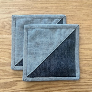May include: Two denim coasters with a diagonal seam. The top coaster is light blue denim and the bottom coaster is dark blue denim.