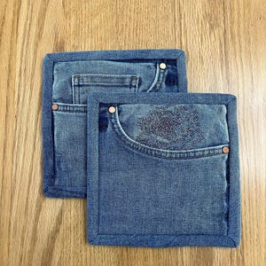 May include: Two square denim pot holders with a blue jean pocket design. One has a floral embroidery on the pocket. Both have copper rivets and a dark blue border. The pot holders are on a wooden surface.