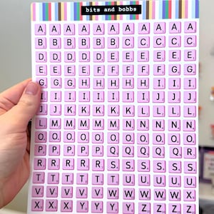 May include: A pink and white Scrabble tile board with the text "bits and bobbs" at the top. The board is filled with letter tiles, perfect for word games. The tiles are pink with white letters and numbers.