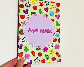 Op de afbeelding: Een lichtgroene notitieboekomslag met een herhalend patroon van kleurrijke aardbeien, knopen, hartjes en sterren. In het midden bevat een lichtpaarse geschulpte cirkel de tekst JUNE PAPER in dikke, roze, afgeronde letters.