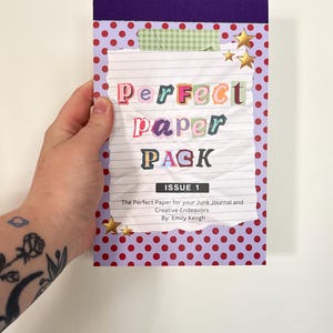 May include: A notepad with a purple spine and a cover featuring the text "Perfect Paper Pack" in colorful letters. The cover has a red polka dot pattern and gold star embellishments. The notepad is designed for junk journals and creative endeavors.