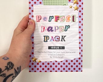 Op de afbeelding: Een notitieblok met een paarse rug en een omslag met de tekst "Perfect Paper Pack" in kleurrijke letters. De omslag heeft een rood stippenpatroon en gouden sterversieringen. Het notitieblok is ontworpen voor junk journals en creatieve inspanningen.