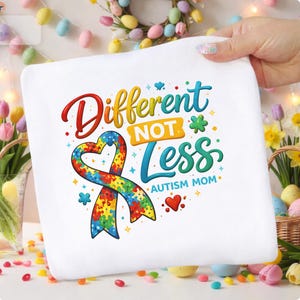 Könnte beinhalten: Weißes quadratisches Kissen mit den Worten "Different Not Less" in farbenfrohen Buchstaben. Ein Band in Herzform befindet sich in der Mitte, hergestellt aus Puzzleteilen. Die Worte "Autism Mom" stehen unten.