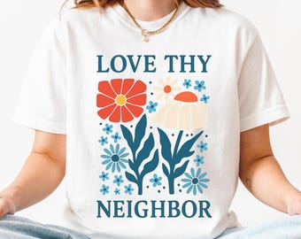 Op de afbeelding: Wit T-shirt met een bloemenpatroon en de tekst "LOVE THY NEIGHBOR" in teal. Het ontwerp bevat een rode bloem met een gele kern, een crème bloem, blauwe bloemen en teal bladeren.