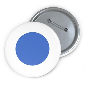 May include: Two white button badges with a blue circle design. One badge is in focus, displaying the front with the blue circle, while the other shows the back with a pin. The badges are likely for clothing or accessories.