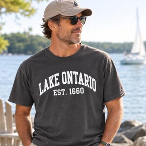 May include: A charcoal gray short-sleeved t-shirt featuring the white text "LAKE ONTARIO EST. 1660" printed across the chest in a bold, arched font. The shirt is made of a soft, washed-out fabric and has a classic crew neckline.