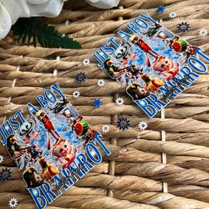 May include: Two diamond-shaped stickers with the text "JUST A BOY BR MAROT who love" in blue. The stickers feature a colorful cartoon design with various characters. The background is a woven, light brown material, with blue star-shaped confetti.