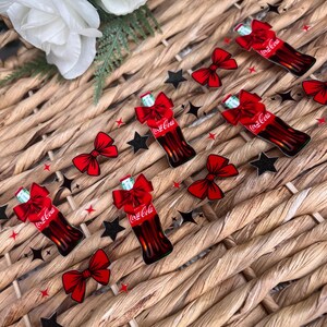 May include: A set of six red and white Coca-Cola bottle charms with bows. The charms are on a woven wicker background.