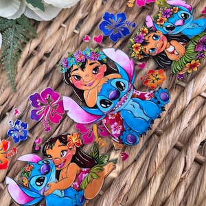 May include: Three colorful stickers featuring Lilo and Stitch from the Disney movie Lilo & Stitch. Each sticker features Lilo and Stitch in a different pose, with Lilo wearing a flower lei and Stitch wearing a flower lei and a pink and white floral shirt. The stickers are on a woven wicker background.
