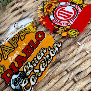 May include: Two colorful stickers, one with a soccer ball and the text "Papa eres un Diablo con buen corazón" and the other with a red and white logo with the text "Club Deportivo Toluca".