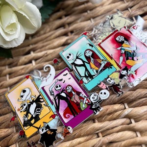 May include: A collection of colorful enamel pins featuring characters from "The Nightmare Before Christmas." The pins showcase Jack Skellington, Sally, and other characters in various scenes. The pins have a glossy finish and are set against a woven background.