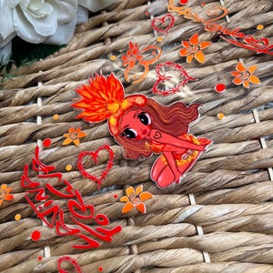 May include: A collection of transparent stickers featuring a cartoon woman with red skin, brown hair, and a floral headdress. The stickers also include orange flowers, hearts, and Arabic script in red. The stickers are arranged on a woven surface.